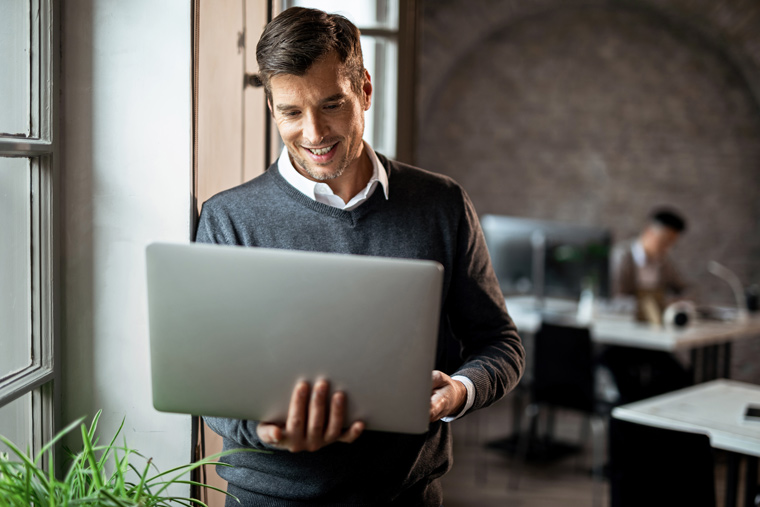 Homem em pé em escritório, utilizando notebook.